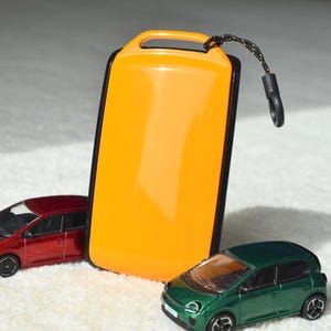 Peut inclure: Un étui rectangulaire orange avec une bordure noire et une lanière noire, placé entre deux voitures miniatures. Une voiture est rouge, l'autre est verte. L'étui a une poignée en haut.