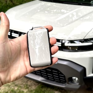Könnte beinhalten: Eine rechteckige Autoschlüssel-Schutzhülle mit glänzendem, hellbeigem Finish und schwarzem Rand. Die Hülle verfügt über eine kleine integrierte Öse an der Oberseite zur Befestigung und bietet eine glatte, reflektierende Oberfläche für den Schlüssel.