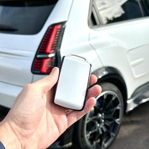 Puede incluir: Un llavero de coche blanco con borde negro se sostiene frente a un SUV blanco. El SUV tiene ruedas negras y un acento azul en el techo. Las luces traseras del coche son rojas.