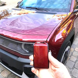 Op de afbeelding: Een bordeauxrode auto met een bijpassende sleutelhanger. De auto heeft een zwarte voorbumper en een reflecterende voorruit. De sleutelhanger wordt voor de auto gehouden, waarbij de kleur en het ontwerp worden getoond.