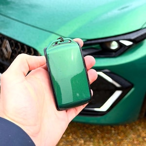 Peut inclure: Un porte-clés de voiture vert émeraude brillant avec une bordure noire. Le porte-clés est tenu devant une voiture verte assortie, avec le phare et le pare-chocs avant de la voiture en arrière-plan.