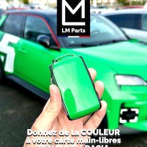 Puede incluir: Una funda de llavero verde brillante con un borde negro se sostiene frente a un coche verde. La funda tiene un acabado brillante. La imagen incluye el texto "LM Parts" y texto en francés que se traduce como "Da color a tu tarjeta manos libres RENAULT / DACIA."