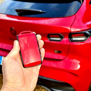 Puede incluir: Un llavero de coche rojo con borde negro. El llavero se sostiene frente a un coche rojo, con la parte trasera del coche en el fondo. El coche tiene detalles en negro y un limpiaparabrisas trasero.