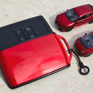 Può includere: Una cover rossa lucida per chiave auto con interno nero e un anello in plastica nera. La cover ha una forma rettangolare con bordi arrotondati e un'apertura per i pulsanti. Due piccole auto giocattolo rosse sono posizionate accanto.