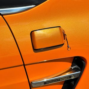 May include: Close-up of an orange car panel with a metallic finish. Features include a fuel door with a black border and a chrome accent with the text "RENAULT SPORT". The car's paint has a shimmering effect.