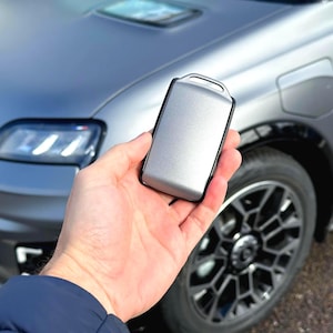 Könnte beinhalten: Ein silberner Autoschlüsselanhänger mit schwarzem Rand wird vor einem grauen Auto gehalten. Das Auto hat ein schlankes Design mit schwarzen Rädern und einem Ladeanschluss. Der Schlüsselanhänger ist rechteckig mit einer Schlaufe.