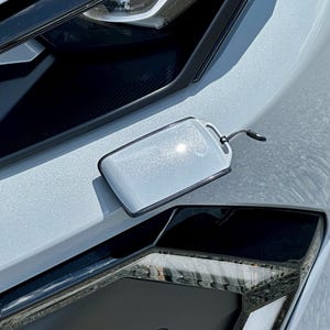 May include: A rectangular, light grey, metallic-finish key fob cover with a smooth, glossy surface and a black trim edge. The cover features a small circular hole at the top right corner with a thin black cord loop attached for secure handling.
