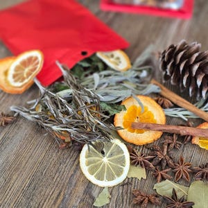 Rosemary, dried oranges, cinnamon sticks from simmer pot contents spread out from a red bag with a pinecone on the side.