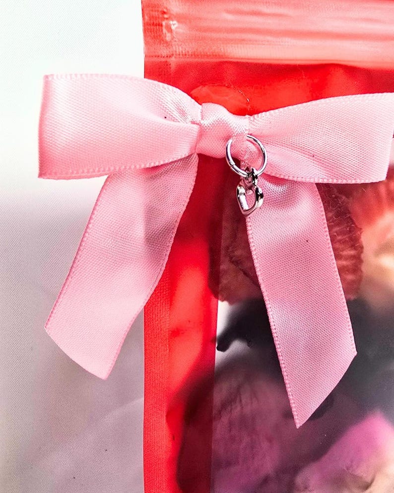 Pu&ograve; includere: Un fiocco di raso rosa adorna un pacco rosso. Un piccolo ciondolo d'argento con un ciondolo a forma di cuore pende dal nodo del fiocco. I nastri del fiocco sono ben disposti, creando un elemento decorativo.