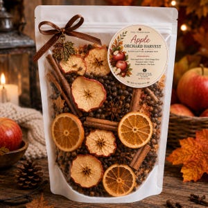 May include: A clear pouch containing a potpourri mix of dried apple slices, orange slices, cinnamon sticks, star anise, and cloves. A brown ribbon with a metal maple leaf charm is tied at the top. A circular label reads Apple Orchard Harvest.