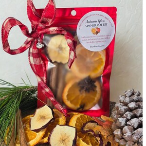 Peut inclure: Un sachet rouge en cellophane étiqueté "Autumn Glow Simmer Pot Kit" rempli de tranches d'orange séchées, de bâtons de cannelle et d'anis étoilé. Le sac est noué avec un ruban à carreaux rouge et blanc et un charm en forme de cœur argenté. Le kit est entouré d'aiguilles de pin, de fruits secs et d'une pomme de pin.