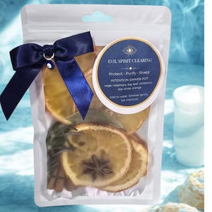 May include: A clear pouch tied with a navy blue ribbon and an evil eye charm. The pouch contains dried orange slices, cinnamon sticks, star anise, and herbs. A circular label reads "EVIL SPIRIT CLEARING" and details the intention simmer pot ingredients.