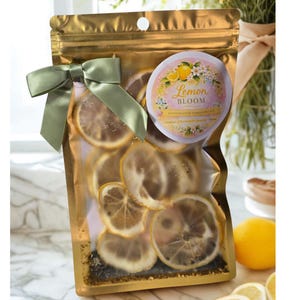 Puede incluir: Una bolsa dorada sellada llena de rodajas de limón secas, atada con una cinta verde salvia. Una etiqueta circular dice "Lemon Bloom" con ilustraciones florales. La bolsa contiene un kit de ducha artesanal.