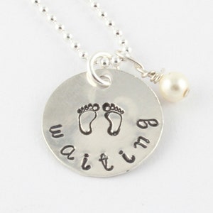 Puede incluir: Collar de plata con un colgante redondo que dice "waiting" con dos huellas de pies de bebé y un pequeño dije de perla.