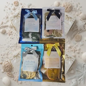 Könnte beinhalten: Vier Potpourri-Beutel mit dekorativen Schleifen. Die Beutel sind blau, transparent und goldfarben, jeweils mit einem Etikett mit der Aufschrift "Salt Air + Sage", "Coconut Coast", "Sea Glass Serenity" und "Golden Hour in the Gulf". Der Hintergrund zeigt Muscheln und kleine Geschenkboxen.