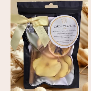 May include: A black pouch containing a simmer pot blend with dried apple slices, orange slices, cinnamon sticks, and other botanicals. The pouch is tied with a yellow ribbon and a small house charm. Text on the label reads "House Blessing".