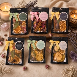 Puede incluir: Seis bolsas negras que contienen botánicos secos, rodajas de naranja y ramas de canela, etiquetadas con temas como Endurance, Self-Love, Joy & Success, Protection, Healing y House Blessing, adornadas con cintas de colores y dijes.