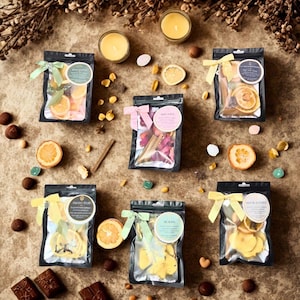 Può includere: Vista dall'alto di diversi piccoli sacchetti trasparenti pieni di frutta secca e prodotti botanici, legati con nastri. I sacchetti sono disposti attorno a candele, fette di frutta secca e cioccolatini su una superficie strutturata. I sacchetti hanno etichette.