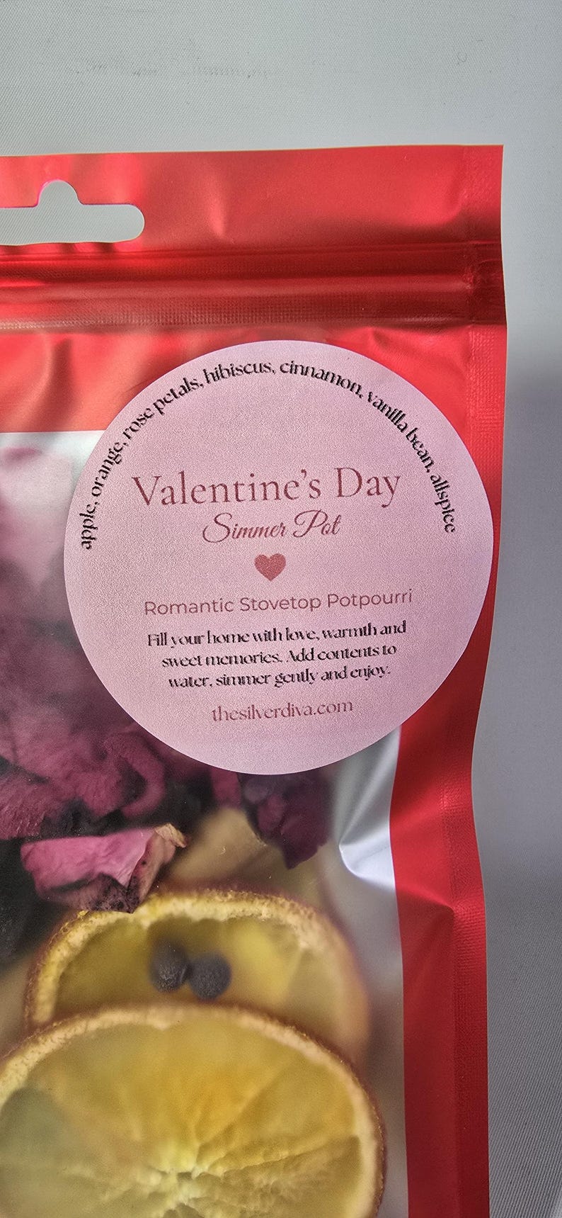 Pu&ograve; includere: Un sacchetto trasparente di Simmer Pot di San Valentino con petali di rosa secchi, fette d'arancia e spezie. L'etichetta recita "Simmer Pot di San Valentino" e "Pot-pourri romantico per fornelli".