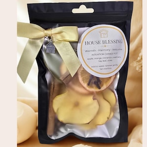 A black pouch containing a simmer pot kit. The kit includes dried apple slices, orange slices, cinnamon sticks, and other botanicals. A yellow ribbon and house charm adorn the pouch. The label reads "House Blessing" and lists ingredients.