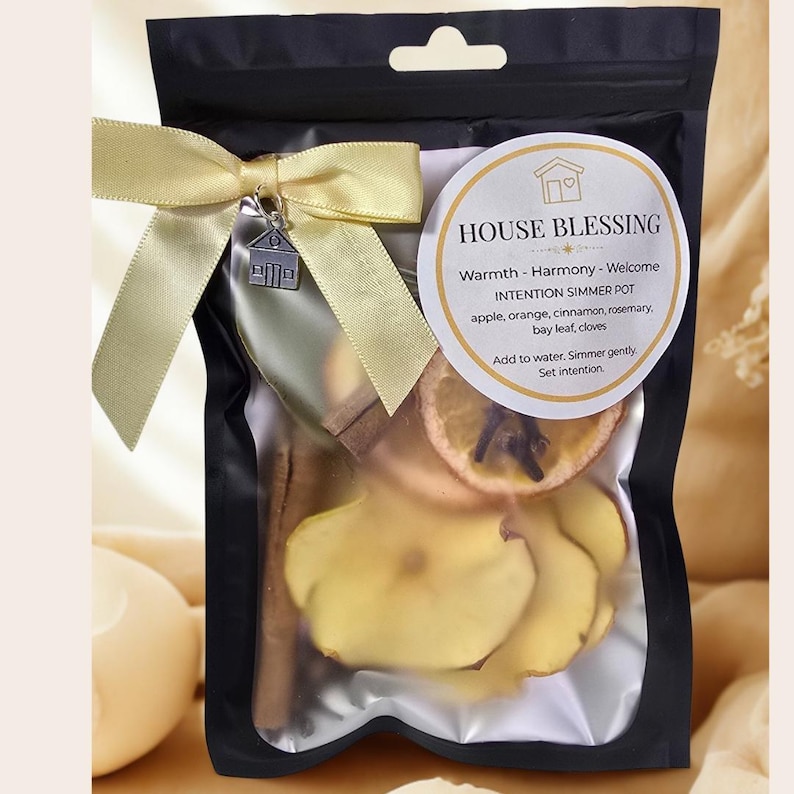 A black pouch containing a simmer pot kit. The kit includes dried apple slices, orange slices, cinnamon sticks, and other botanicals. A yellow ribbon and house charm adorn the pouch. The label reads "House Blessing" and lists ingredients.