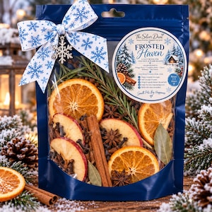 May include: A dark blue pouch containing a simmer pot kit labeled Frosted Haven. The kit includes dried orange slices, apple slices, cinnamon sticks, star anise, cloves, rosemary, and bay leaves. A white ribbon with blue snowflakes and a snowflake charm is attached.