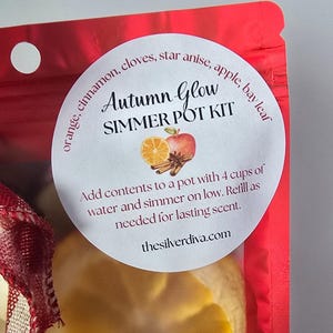 May include: A red package with a white label that reads "Autumn Glow Simmer Pot Kit." The label features illustrations of an apple, orange slice, and cinnamon sticks. The text on the label also includes instructions and the website thesilverdiva.com.