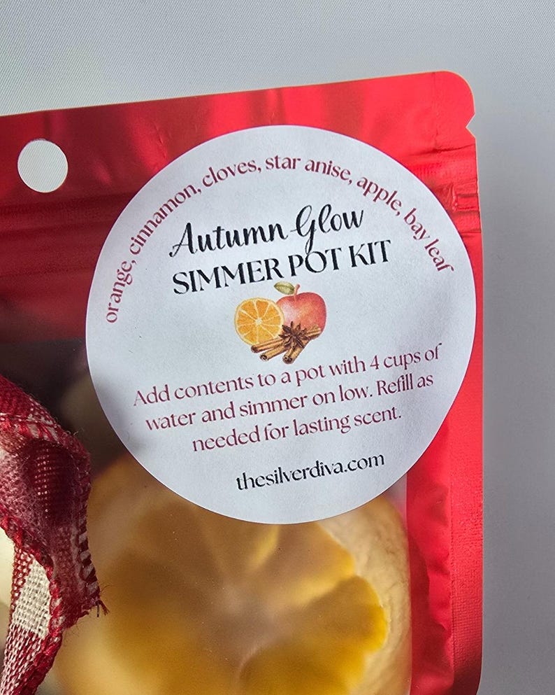May include: A red package with a white label that reads "Autumn Glow Simmer Pot Kit." The label features illustrations of an apple, orange slice, and cinnamon sticks. The text on the label also includes instructions and the website thesilverdiva.com.