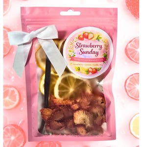 Puede incluir: Una bolsa rosa con rodajas de fruta deshidratada, una vaina de vainilla y un lazo blanco. Una etiqueta circular dice "Strawberry Sunday Handcrafted Simmer Pot Kit" con ingredientes de fresa, limón, vainilla y menta.