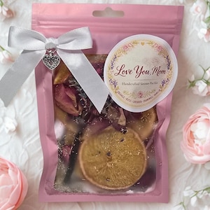 Puede incluir: Una bolsa rosa atada con una cinta blanca y un dije de corazón, que contiene flores secas y una rodaja de naranja. Una pegatina redonda dice "Love You, Mom" con detalles florales. Es un kit de olla a fuego lento hecho a mano.