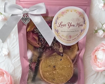 May include: A pink pouch tied with a white ribbon and a heart charm, containing dried flowers and a sliced orange. A round sticker reads "Love You, Mom" with floral accents. The pouch is a handcrafted simmer pot kit.