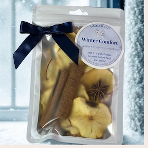 May include: A clear plastic bag tied with a navy blue bow, containing dried apple slices, cinnamon sticks, and star anise. A circular label reads "Simmer Pot Winter Comfort" with instructions. The ingredients are for a warm, cozy, and comforting simmer pot.