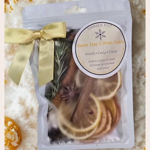 May include: A clear plastic bag tied with a gold ribbon, containing dried citrus slices, cinnamon sticks, and herbs. The bag has a circular label that reads "Snow Day Citrus Spice". The label also says "Fresh, Cozy, Clean".