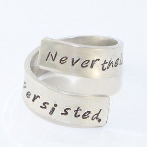 Puede incluir: Un anillo envolvente plateado con las palabras "Never the" y "Persisted" grabadas en la superficie. El anillo tiene un diseño moderno y minimalista y está sobre un fondo blanco.