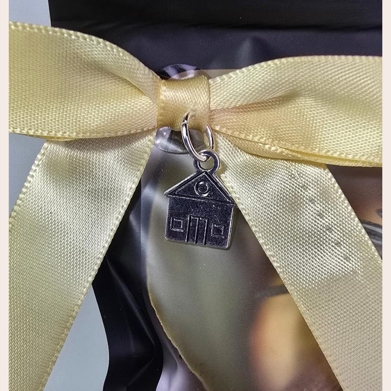 House blessing simmer pot gift bag tied with a pale yellow satin ribbon and bow. A small silver house charm hangs from the bow's knot. The bag is black and the background is a light beige.