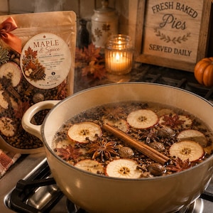 May include: A beige Dutch oven on a hob filled with simmering water, dried apple slices, cinnamon sticks, star anise, and cloves. Beside it sits a matching pouch labelled Maple & Spice with a burnt orange ribbon and a maple leaf charm.