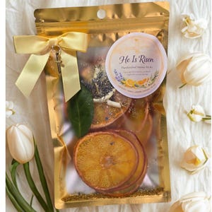 Puede incluir: Una bolsa dorada atada con una cinta amarilla y un dije de cruz. Contiene rodajas de naranja deshidratadas, hierbas y una etiqueta que dice "He Is Risen" y "Handcrafted Simmer Pot Kit".