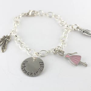 Pode incluir: Pulseira de prata com um berloque de prata que diz "Quebra-Nozes 2017", um berloque de prata de uma bailarina, um berloque de prata de um par de sapatilhas de balé e um berloque de prata de um quebra-nozes.