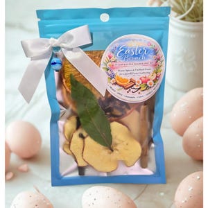 Puede incluir: Una bolsa azul atada con una cinta blanca y una cuenta azul y blanca. La bolsa contiene fruta seca, especias y una hoja verde. Una pegatina redonda en la bolsa dice "Easter Brunch" y enumera los ingredientes. Huevos de Pascua rosas moteados están en el fondo.