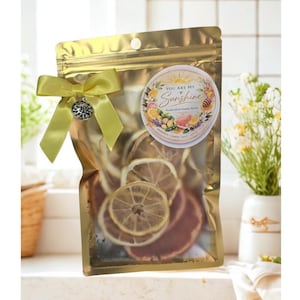 Puede incluir: Una bolsa dorada llena de rodajas de cítricos secos, atada con una cinta amarilla y un dije plateado. Una pegatina circular en la bolsa dice "You Are My Sunshine" con ilustraciones de frutas. La bolsa es un kit de maceta de verano artesanal.
