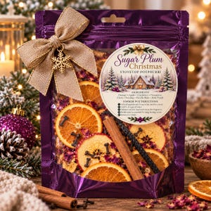 May include: A purple pouch of Sugar Plum Christmas stovetop potpourri featuring dried orange slices, apple slices, cinnamon sticks, and cloves. A tan burlap bow with a gold snowflake charm is attached to the top. The label lists ingredients and directions.