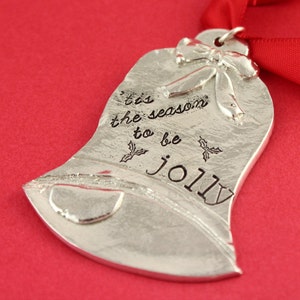 May include: Silver Christmas ornament shaped like a bell with the text 'tis the season to be jolly' and a small bow on top.