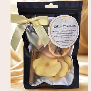 House blessing black pouch containing a simmer pot kit. The kit includes dried apple slices, orange slices, cinnamon sticks, and other herbs. A gold ribbon and a house charm adorn the pouch. The label reads "HOUSE BLESSING" and lists ingredients.