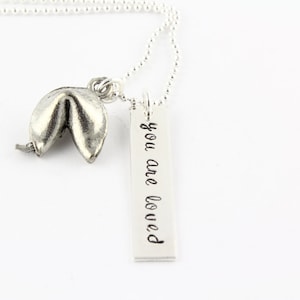 Pode incluir: Colar de prata com um pingente de biscoito da sorte e uma etiqueta que diz "you are loved".