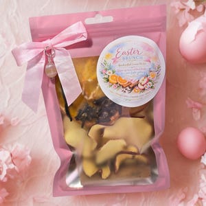 Puede incluir: Una bolsa de celofán rosa atada con una cinta rosa y un dije, llena de frutas secas y especias. Una pegatina redonda en la bolsa dice "Easter Brunch" con ilustraciones florales y frutales. Dos huevos de Pascua rosas en el fondo.