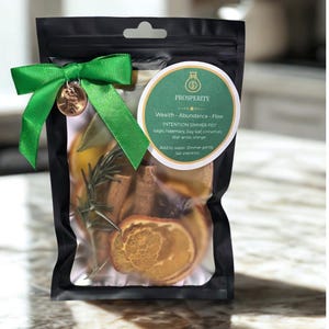 Könnte beinhalten: Ein schwarzer Beutel, gebunden mit einem grünen Band und einem Penny-Anhänger. Der Beutel enthält getrocknete Kräuter und Orangenscheiben. Ein rundes Etikett trägt die Aufschrift "PROSPERITY" und listet die Zutaten auf.