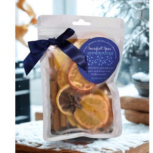 May include: A clear cellophane bag tied with a navy blue ribbon, containing dried orange slices, cinnamon sticks, and star anise. A blue label reads "Snowfall Spice Simmer Pot Kit". The bag is on a wooden surface.