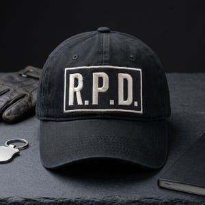 Op de afbeelding: Zwarte baseballcap met de letters "R.P.D." in wit geborduurd in een wit rechthoek. De cap is gemaakt van een donkere stof en heeft een gebogen klep. De cap ligt op een donkere ondergrond.