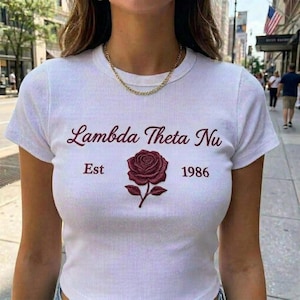 Könnte beinhalten: Weißes geripptes T-Shirt mit dem Text "Lambda Theta Nu" über einer bordeauxfarbenen Rosengrafik. Der Text "Est 1986" befindet sich unter der Rose. Eine goldene Halskette wird getragen.