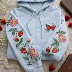 以下が含まれることがあります： 袖と胸にイチゴとピンクの花が刺繍された、水色のフード付きスウェットシャツ。フード付きスウェットシャツには、ドローストリングフードが付いています。新鮮なイチゴがスウェットシャツの周りに散らばっています。